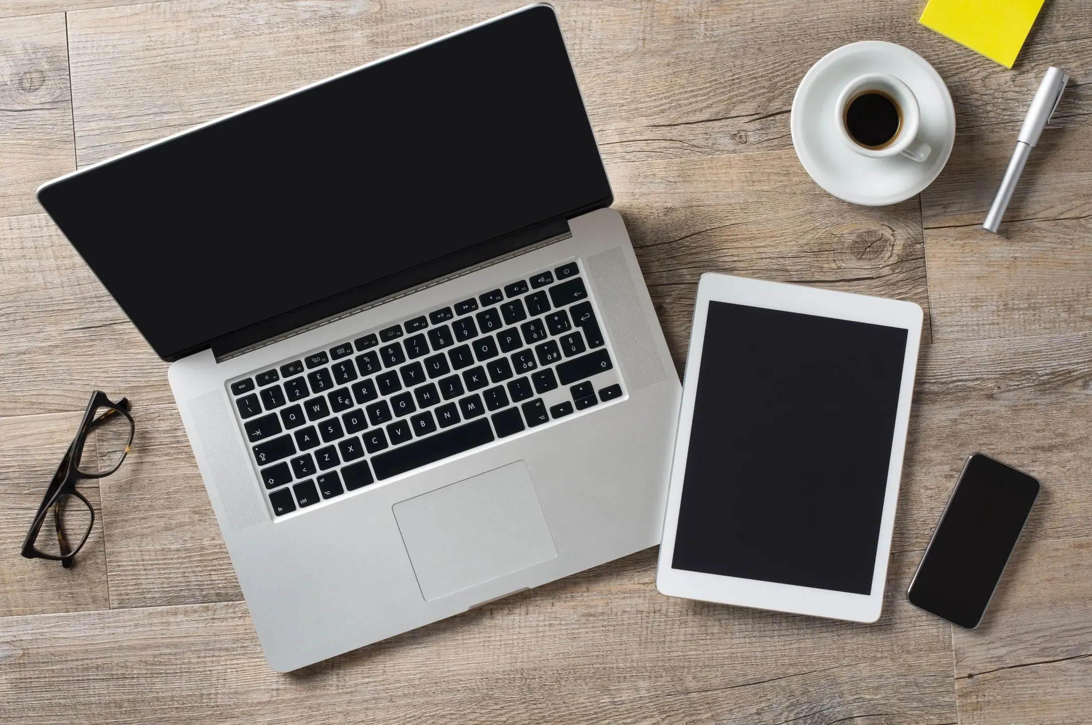 A laptop and tablet side by side on a wooden table with a coffee cup.